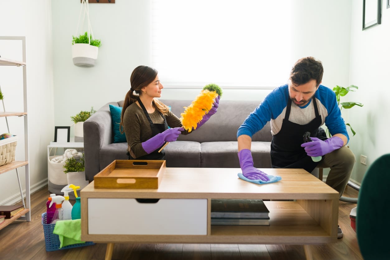 A woman and a man kneeling on the floor behind a coffee table in a family room dusting and polishing the furniture.